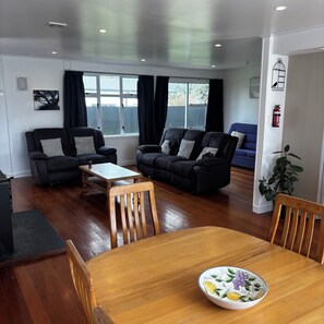 A modern living room with two black sofas, a wooden dining table, and a blue sofa in the background. - The Bach on Dorset 🌊 (Opunake)