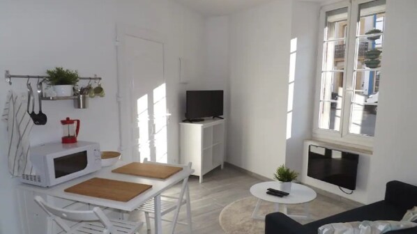 A modern, well-lit living room with a flat-screen TV, a dining table with chairs, a small round table with a plant, and a black sofa.