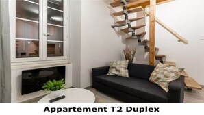 A living room with a black sofa, two patterned cushions, a small table with a plant and a remote control, a fireplace, and a staircase with wooden steps. - DUPLEX DU GET ★ CENTER ★ WIFI (Revel)