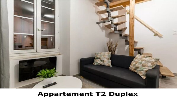 A living room with a black sofa, two patterned cushions, a small table with a plant and a remote control, a fireplace, and a staircase with wooden steps.