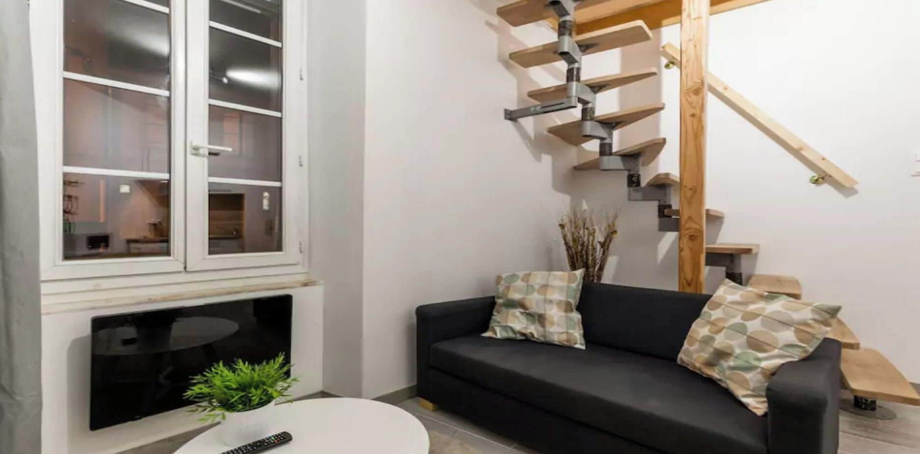 Un salon avec un canapé noir, une table basse blanche, un téléviseur et un escalier en colimaçon.