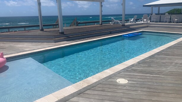 Uma piscina com um deck de madeira, uma pérgula branca e vista para o oceano.