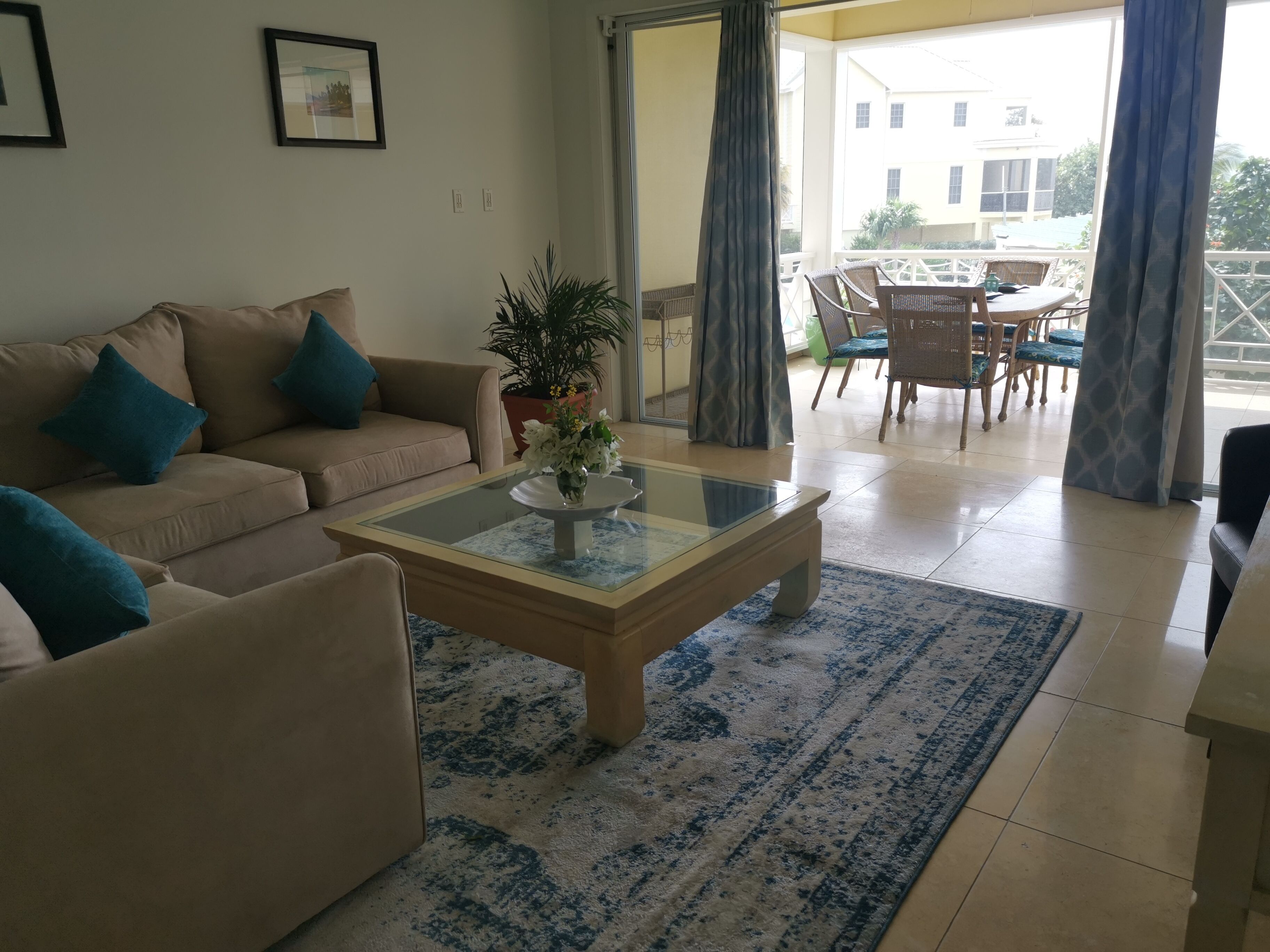 Un salon avec un canapé beige, une table basse en verre ornée d’un vase de fleurs, un tapis à motifs et une vue sur le balcon extérieur.