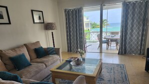 A living room with a beige sofa, a glass coffee table, and a view of the ocean through large windows. - Silver Sand Villa 7A (Cotton Ground)
