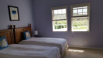A room with two beds, a wooden headboard, a framed picture on the wall, and two windows with a view of greenery.