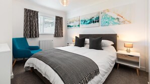 A hotel room with a large bed, a blue armchair, a bedside table with a lamp, and abstract wall art. - 25 Coedrath Park - Sea Views From Balcony Short Walk to Beach Parking (Saundersfoot)