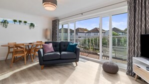 A modern living room with a sofa, wooden dining table, and chairs. There is a large window with a view of houses and a balcony. - 25 Coedrath Park - Sea Views From Balcony Short Walk to Beach Parking (Saundersfoot)