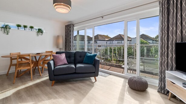 A modern living room with a sofa, wooden dining table, and chairs. There is a large window with a view of houses and a balcony. - 25 Coedrath Park - Sea Views From Balcony Short Walk to Beach Parking (Saundersfoot)