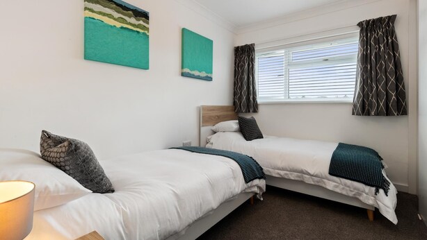 A bedroom with two single beds, a window with curtains, and two wall art pieces. - 25 Coedrath Park - Sea Views From Balcony Short Walk to Beach Parking (Saundersfoot)