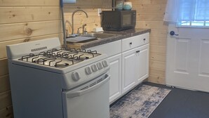 A compact kitchen with a white stove, a microwave, and a sink. There are wooden walls and a window with curtains. - Newfound Lake Cozy Cottage (Bristol)