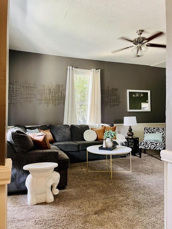 A living room with a dark grey sofa, a white elephant-shaped stool, a round coffee table, and a ceiling fan. - Leaning House of Haynie | Trendy 2BR/1BA Apartment (Evansville)
