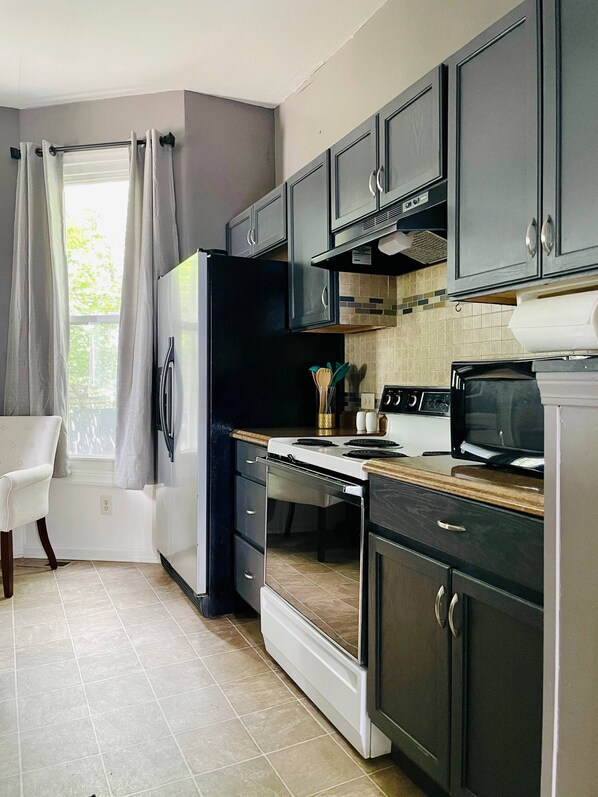 A kitchen with dark cabinets, a refrigerator, an oven, a microwave, and a window with curtains.
