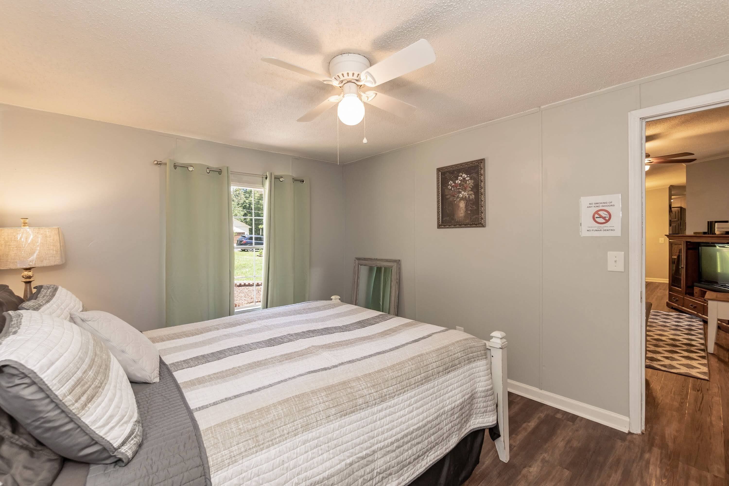 A bedroom with a bed, a ceiling fan, a window with curtains, a door leading to another room, and a small framed picture on the wall.