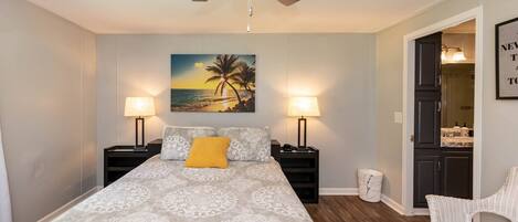 A bedroom with a bed, two bedside tables, a ceiling fan, a night lamp, a wall-mounted painting, and a white chair.