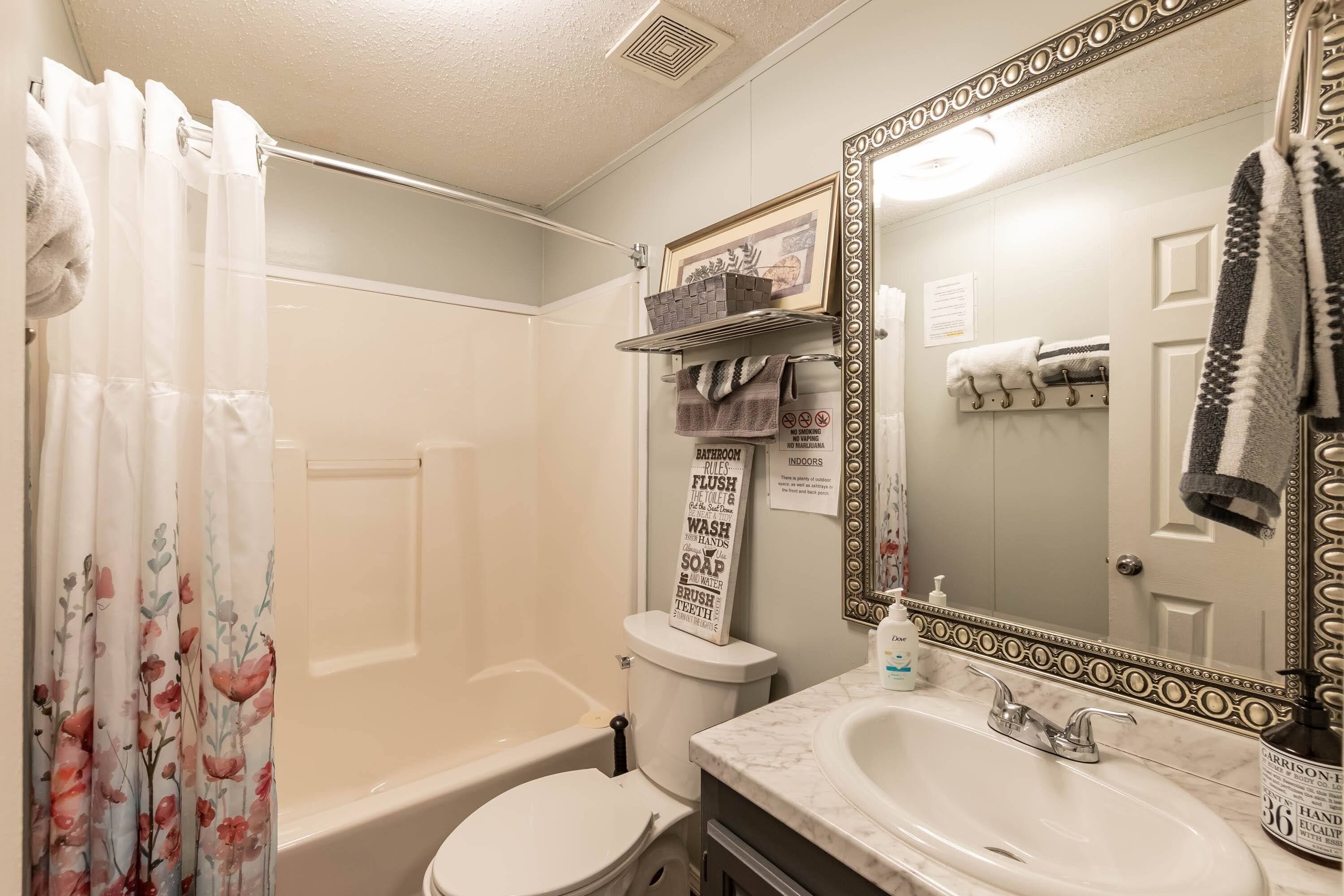 A bathroom with a bathtub, toilet, and sink. There is a shower curtain with floral patterns, a mirror with a decorative frame, and a shelf with towels.