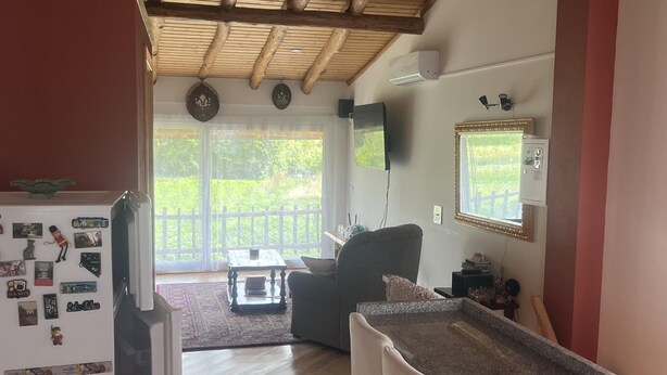 A kitchen with wooden flooring, a refrigerator, a dining area with chairs, a table, and a large window with a view of greenery.
