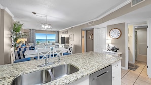 A modern kitchen with a granite countertop, stainless steel sink, and white cabinetry. There is a dining area with a table and chairs, and a view of the ocean through large windows. - Gulf Shores Plantation-Palms 6402 (Gulf Shores)