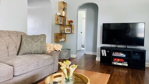 Un soggiorno con un divano grigio, un tavolino in legno con un vaso di fiori, una TV su un mobile nero e una libreria con diversi oggetti.