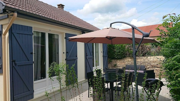 Un patio con una mesa y sillas, un paraguas grande y una casa con contraventanas azules y techo de tejas.