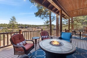 A covered outdoor deck with wicker chairs, a round table with a fire pit, and a view of a forested landscape. - 5 🌟 Luxury Torreon  (Show Low)