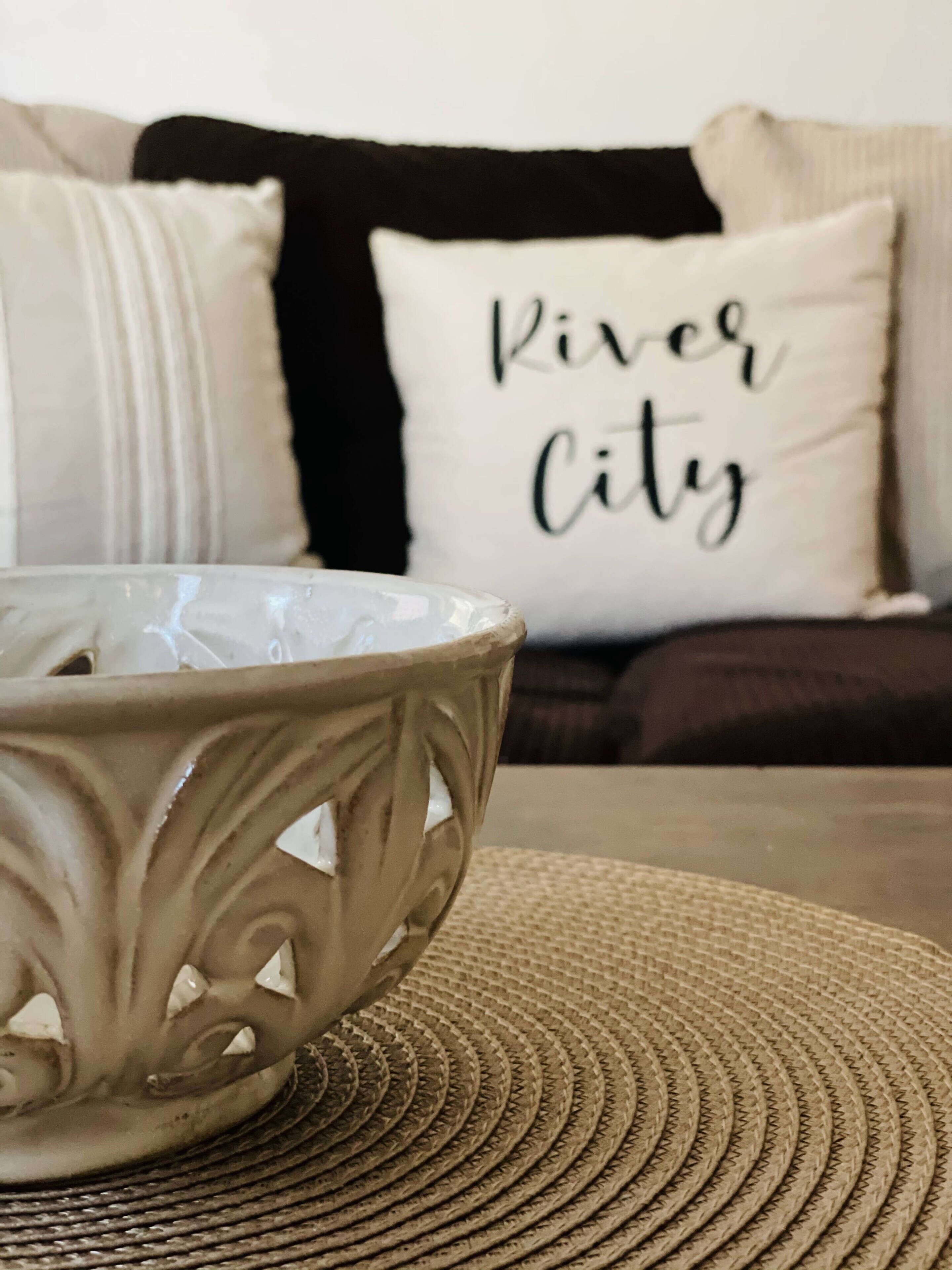 Un bol posé sur un set de table en fibres tressées, un canapé avec un coussin orné de l’inscription « River City », et un mur d’où l’on aperçoit une partie d’une fenêtre.
