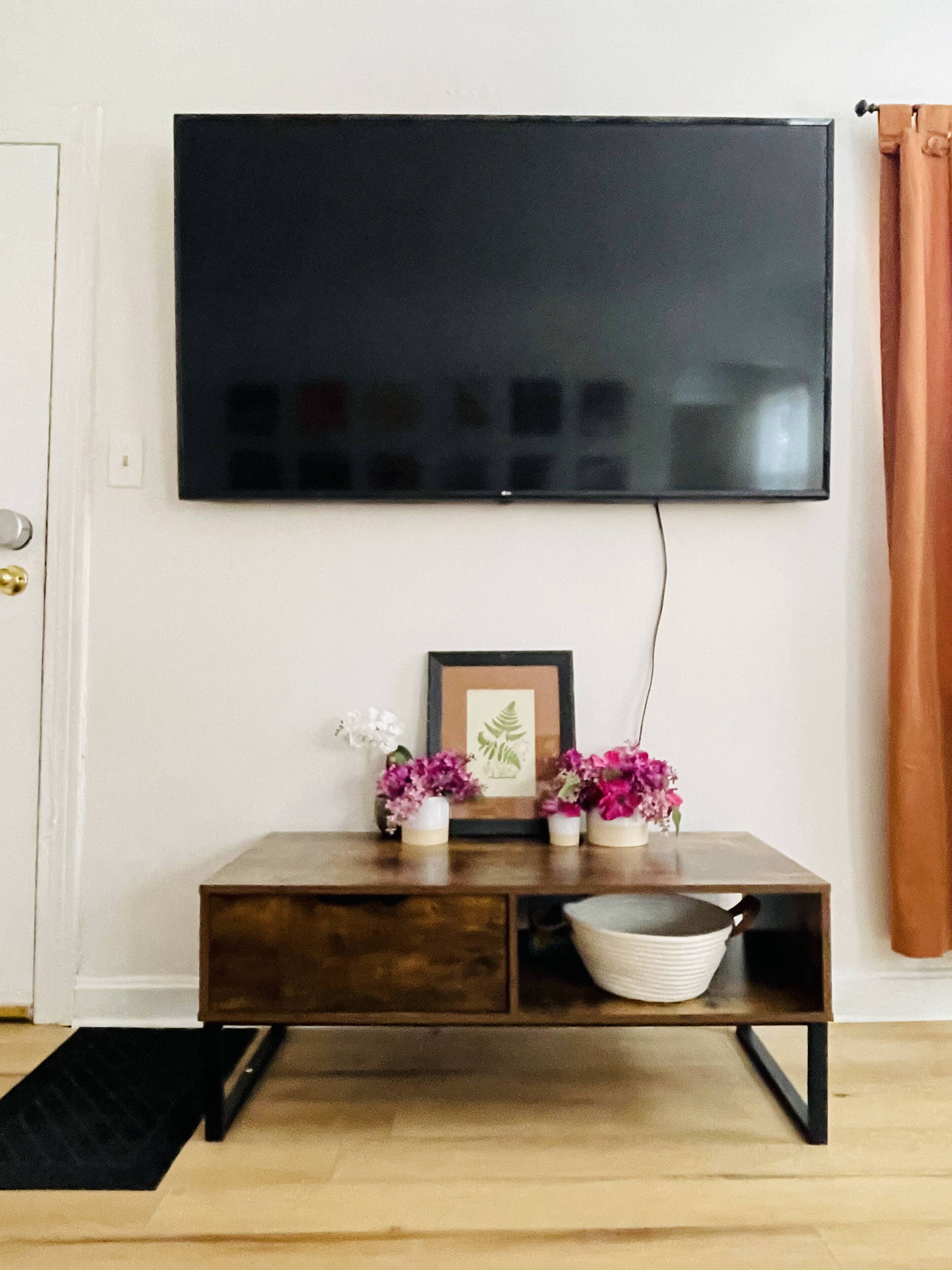 Ett vardagsrum med en platt-TV som är monterad på väggen, ett träbord med dekorativa föremål och en svart dörrmatta.