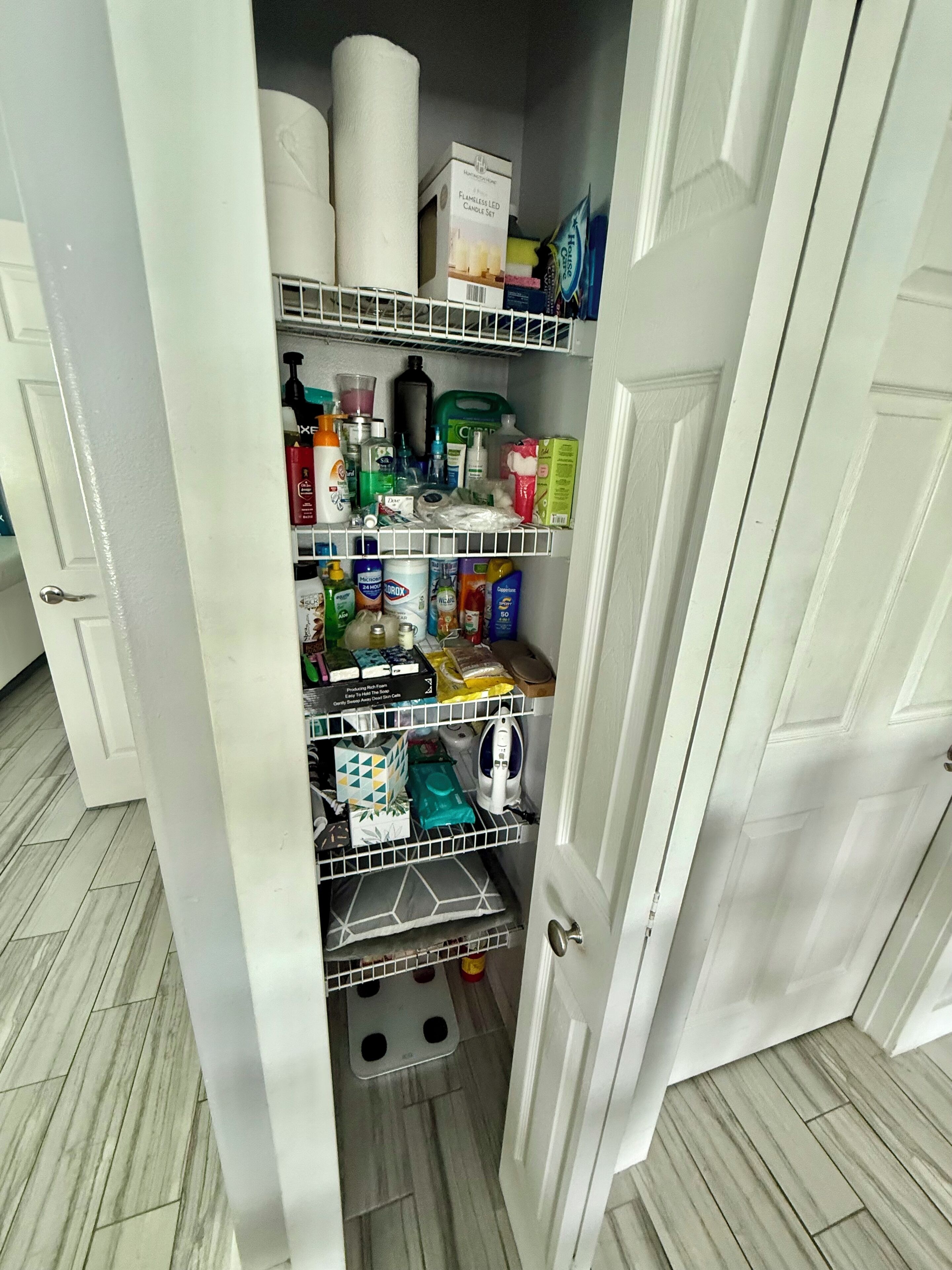 Una piccola e stretta dispensa con scaffali che contengono diversi articoli domestici, come prodotti per la pulizia, candele e articoli da toilette.