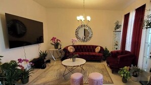 En stue med sofa, skamler, sofabord, fjernsyn, lysekrone og flere planter.