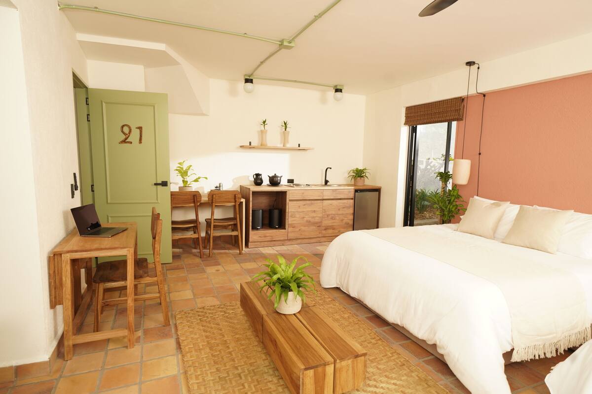 Ein Schlafzimmer mit einem Bett, einem Schreibtisch mit einem Laptop, einer Holzbank mit einer Pflanze und einer Küchenzeile mit Spüle und Schränken.