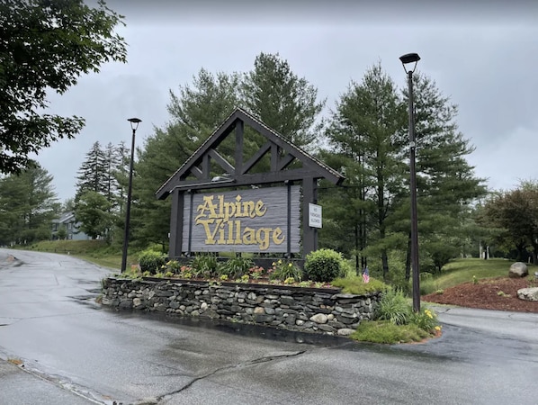 Une enseigne indiquant l’entrée du village alpin, avec un mur de pierres et des plantes devant.