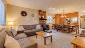Ein Wohnzimmer mit einer Couch, einem Couchtisch und einem Kamin. Es gibt einen Essbereich mit Holzmöbeln und eine Küche im Hintergrund.