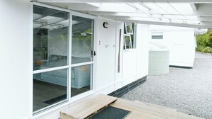 Un edificio moderno con un amplio ventanal corredero, un pequeño porche con terraza de madera y fachada blanca.
