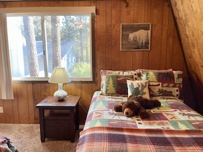 Une chambre chaleureuse avec une grande fenêtre, une tête de lit en bois agrémentée d’oreillers à motifs animaliers, une lampe de chevet et un ours en peluche sur le lit.