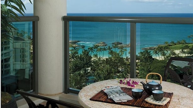 Une table ronde avec un journal, une théière et des tasses, placée sur un balcon donnant sur une plage bordée de palmiers et sur l’océan.