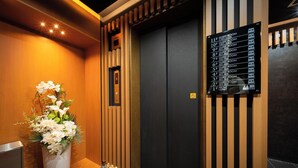 A hotel lobby with a reception desk, a flower arrangement, a black elevator with a guest room directory, and a wooden wall design. - WELLSTAY Shinsekai Wakyo (Osaka)