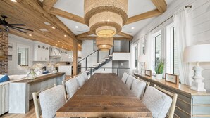Un comedor amplio con una larga mesa de madera rodeada de sillas con acolchado, una lámpara colgante de fibra natural y una escalera que sube a un nivel superior.