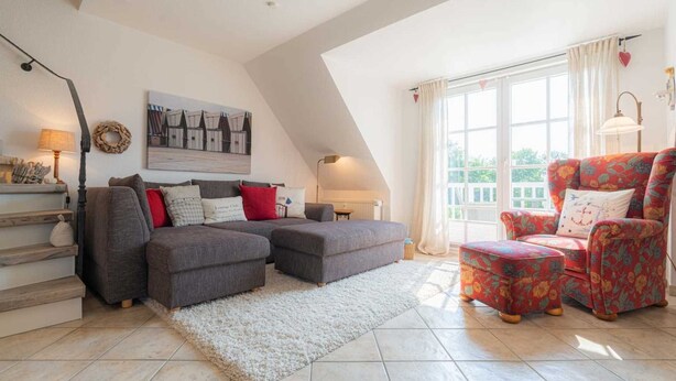 A spacious living room with a grey sectional sofa, a red floral armchair, and a white shag rug. A large window provides natural light, and a painting of boats hangs on the wall. - Apartment 4 Müller (ID 425) (Sankt Peter-Ording)