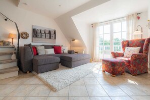 Uma sala de estar ampla com um sofá sectional cinza, uma poltrona vermelha com estampa floral e um tapete felpudo branco. Uma janela grande oferece luz natural e um quadro de barcos está pendurado na parede.