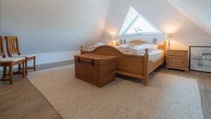 A bedroom with a sloped ceiling, a wooden bed, a small bedside table, a nightstand, a wicker basket, and two chairs. - Apartment 4 Müller (ID 425) (Sankt Peter-Ording)