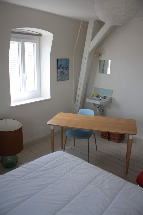 A bedroom with a bed, a wooden table, a blue chair, a window, a sink, and a painting on the wall.