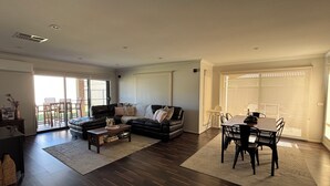 Un vaste salon doté d’un parquet en bois foncé, d’un canapé en cuir noir, d’une table à manger avec des chaises et d’une porte-fenêtre coulissante donnant sur un balcon.