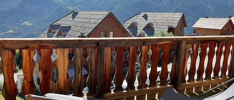 A wooden balcony with a view of mountain houses and a grassy lawn.