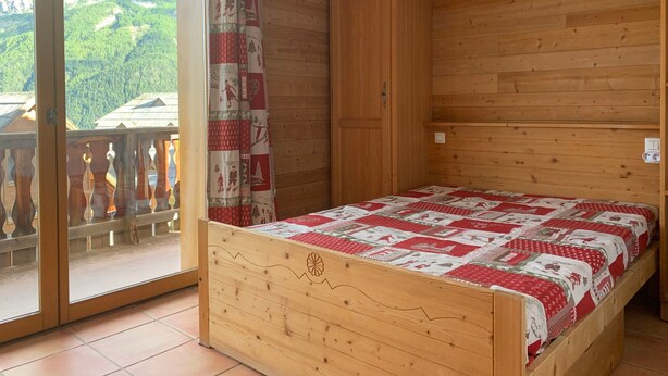Ein Schlafzimmer aus Holz mit Bett, einer Schiebetür zu einem Balkon und Blick auf eine gebirgige Landschaft.