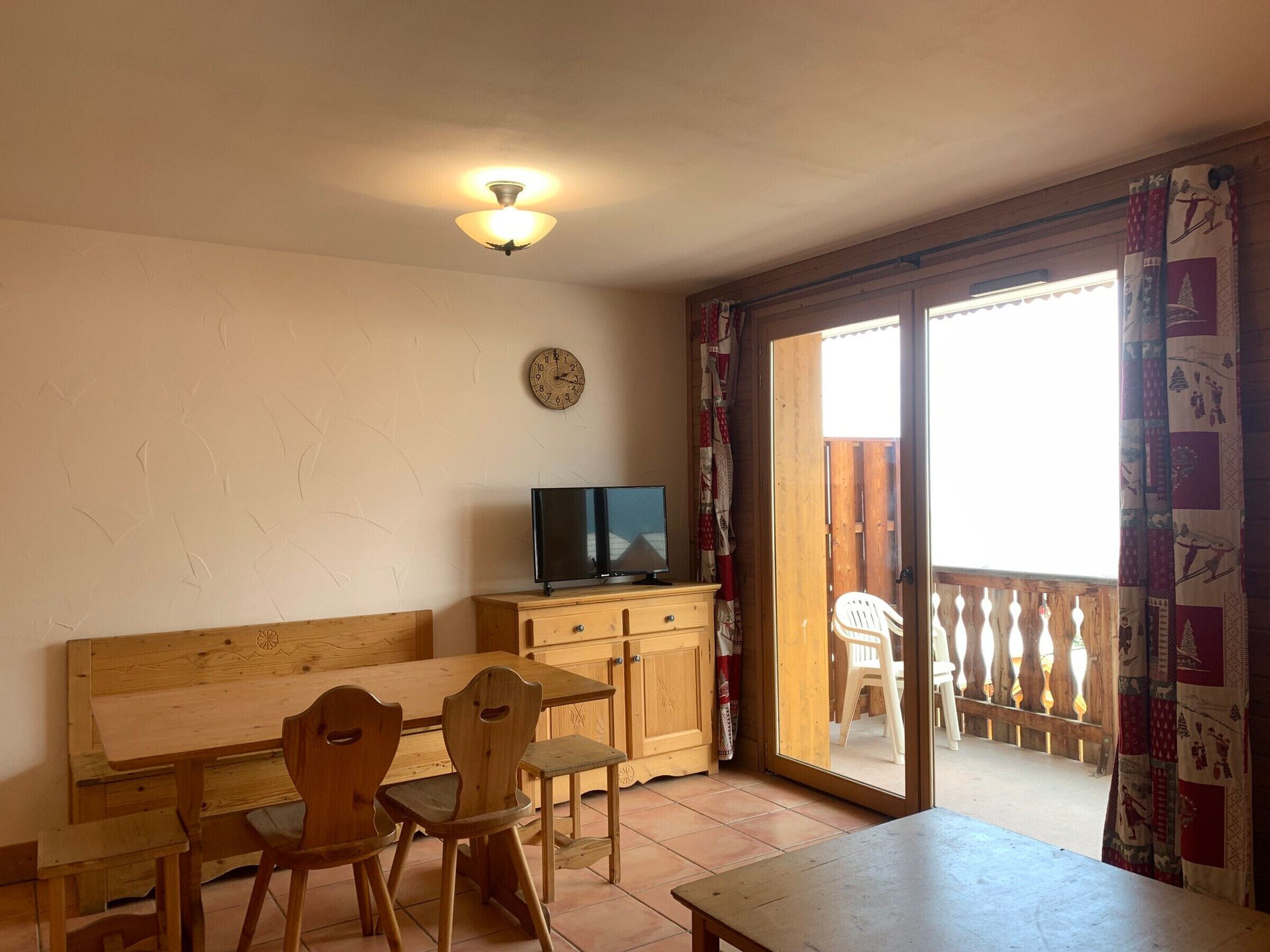 Une pièce chaleureuse avec une table en bois, des chaises, un téléviseur posé sur un meuble, une horloge au mur et une porte-fenêtre coulissante donnant sur un espace extérieur.