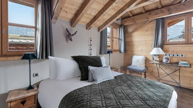A cozy bedroom with a wooden ceiling, a bed with a green quilt, a mounted animal head, and a desk with a lamp and books.