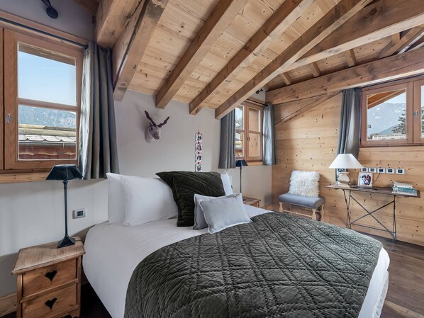 Ein gemütliches Schlafzimmer mit Holzbalkendecke, einem Bett mit grüner Steudecke, einem aufgehängten Tierkopf und einem Schreibtisch mit Lampe und Büchern.