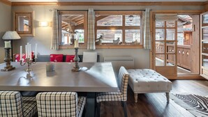 A dining area with a wooden table, checkered chairs, a bench, and a view of the outside through large windows.