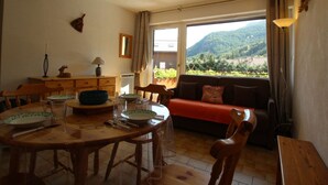 Ein Essbereich mit Holztisch und -stühlen, einer Couch mit rotem Kissen und Blick auf eine Berglandschaft durch ein großes Fenster.