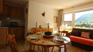 Ein gemütliches Wohnzimmer mit einem Holzesstisch, Stühlen und einer Couch. Im Hintergrund ist eine Küche mit Holzschränken und einer Mikrowelle zu sehen.