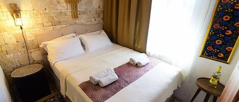 Una cama bien tendida con sábanas blancas y una colcha decorativa roja y blanca, una mesita redonda con una botella, y una alfombra estampada sobre un piso de baldosas.